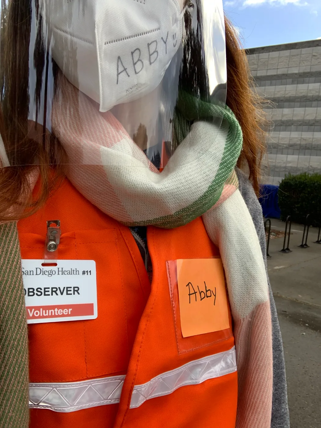 Abby volunteering at a COVID-19 vaccine site in an orange vest with a UC San Diego Health Observer badge and a mask with ABBY written on it